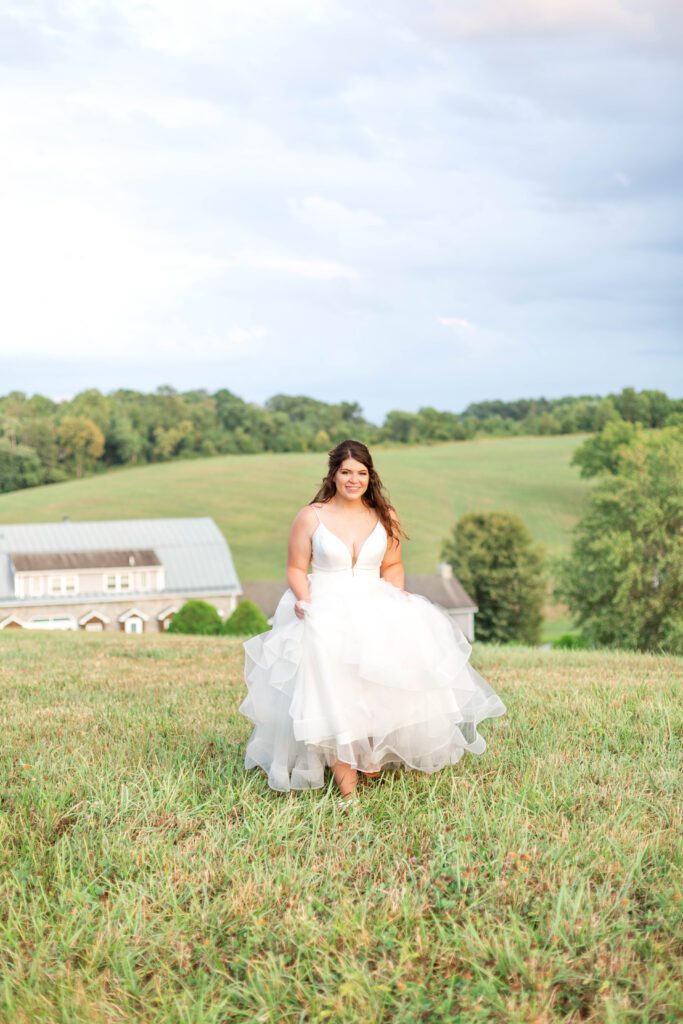 A smiling person in a white wedding dress standing on a grassy field with a house and greenery in the background.