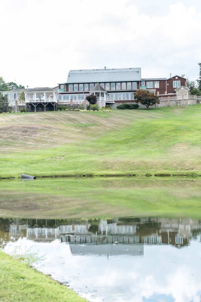 A large country house with a glass-fronted upper level and a wooden lower level, raised on stilts, overlooking a calm pond with the house's reflection visible in the water.