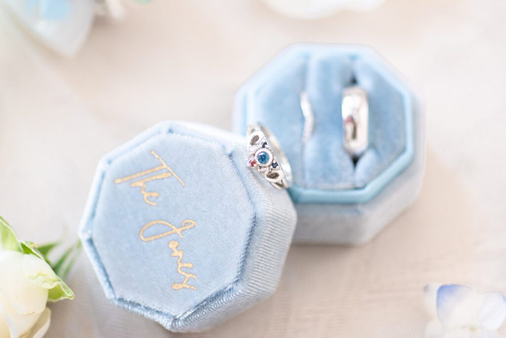 Two blue velvet ring boxes, one with the lid open displaying a silver ring with a blue gemstone and the other closed with "The Jones" embroidered in gold script on the top, placed on a light background with blurred flowers in the foreground.