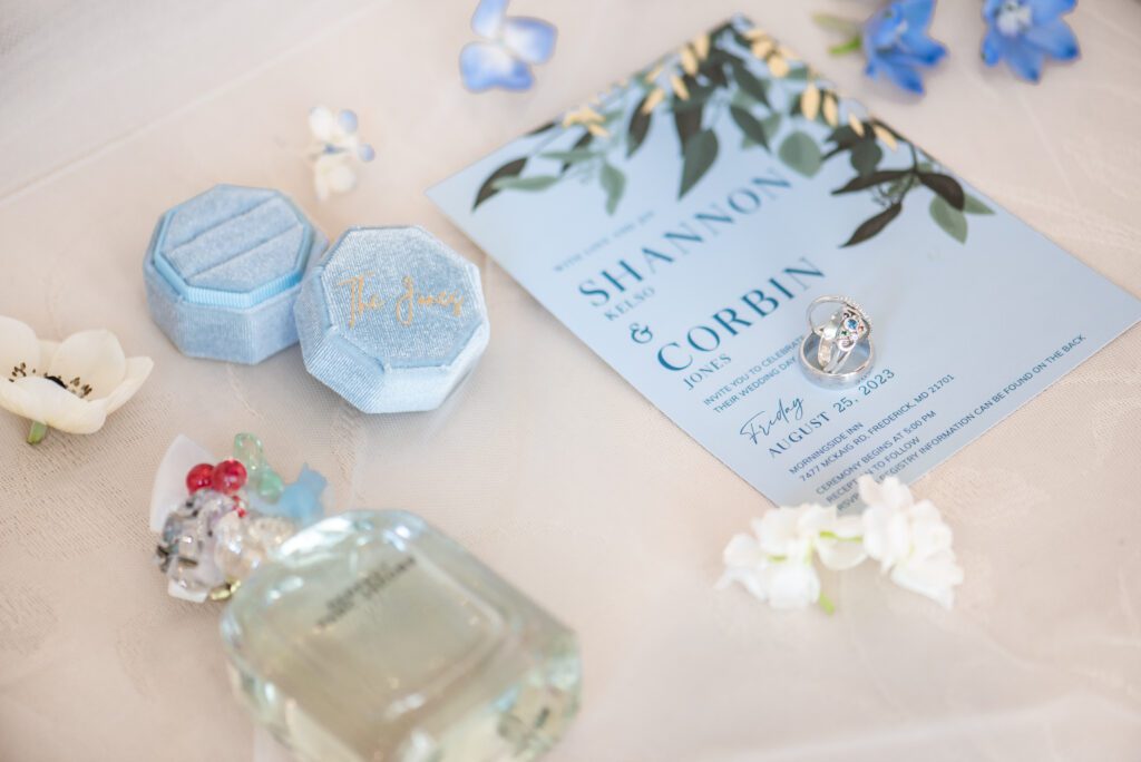 Alt text: A wedding invitation card featuring the names "SHANNON & CORBIN" with a date marked for August 25, 2023, lies beside an open blue ring box with "The Rings" embroidered inside the lid and an engagement ring staged on top of the card. Additional items include a bottle of perfume, scattered flower blossoms, and a small, colorful figurine, all arranged on a textured tablecloth.