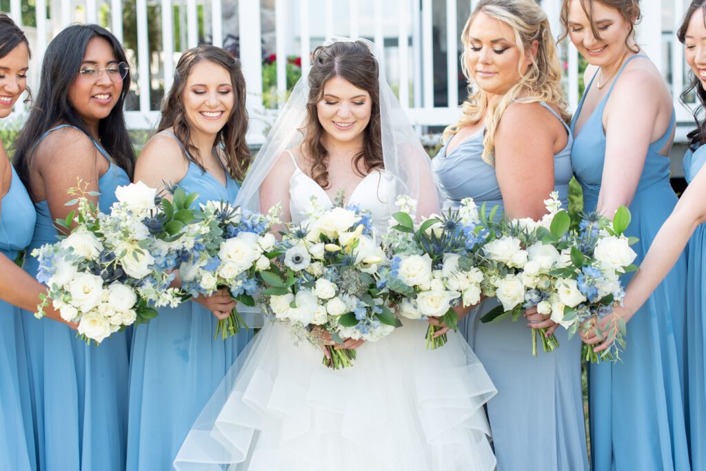 A bride in a white dress and veil surrounded by bridesmaids dressed in light blue, all holding bouquets of white and blue flowers.