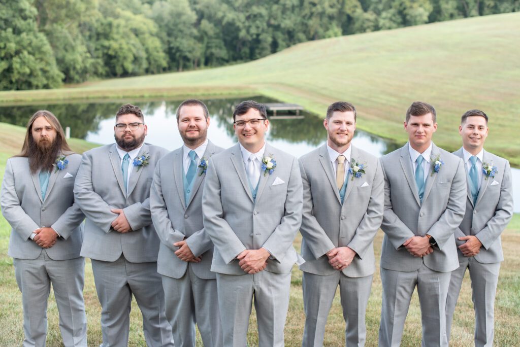 A group of seven men in matching light gray suits and blue boutonnieres standing in a line outdoors, with a calm pond and greenery in the background.