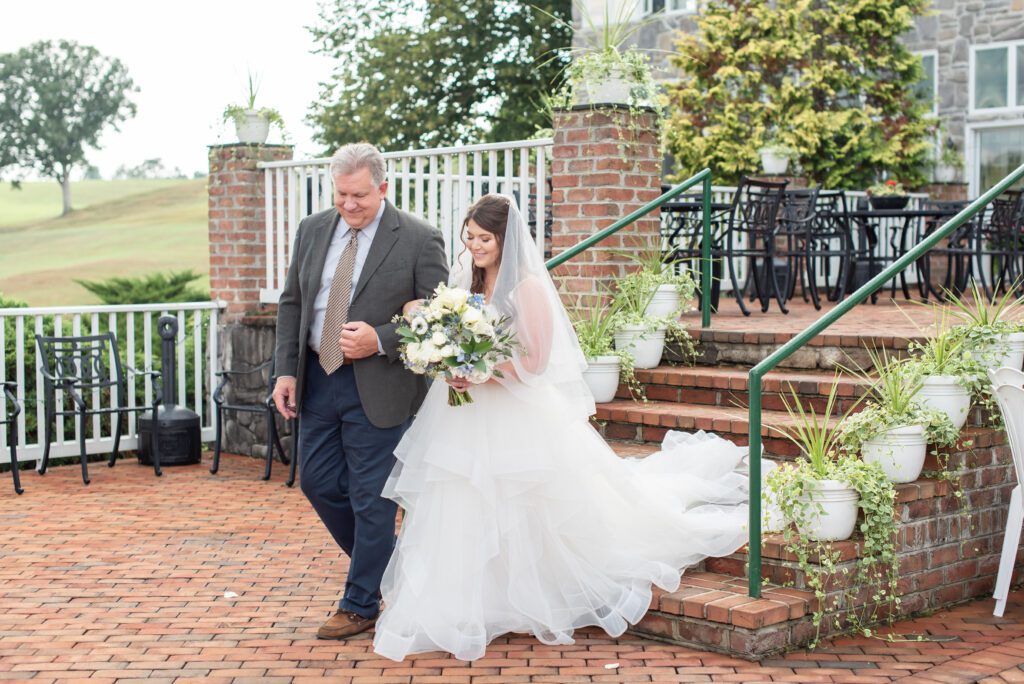 A bride in a white dress and veil holding a bouquet walks down brick steps beside an older man in a suit, with patio furniture and greenery in the background.