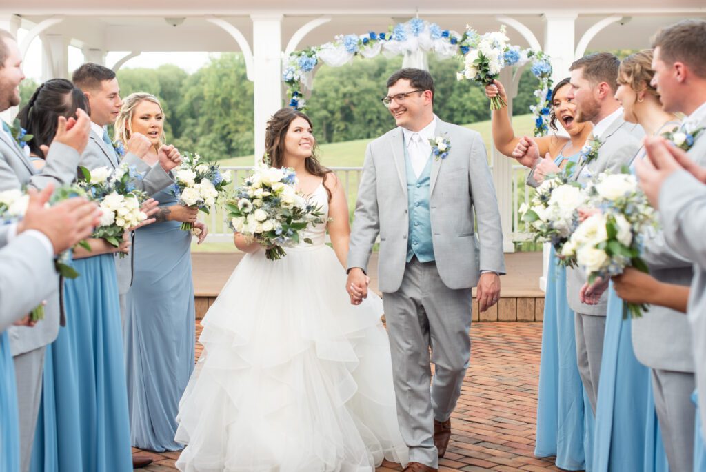 A bride and groom holding hands and smiling at each other as they walk through a cheering bridal party, with attendees wearing blue and holding bouquets. They are under a white gazebo adorned with blue and white flowers, suggesting a wedding celebration.