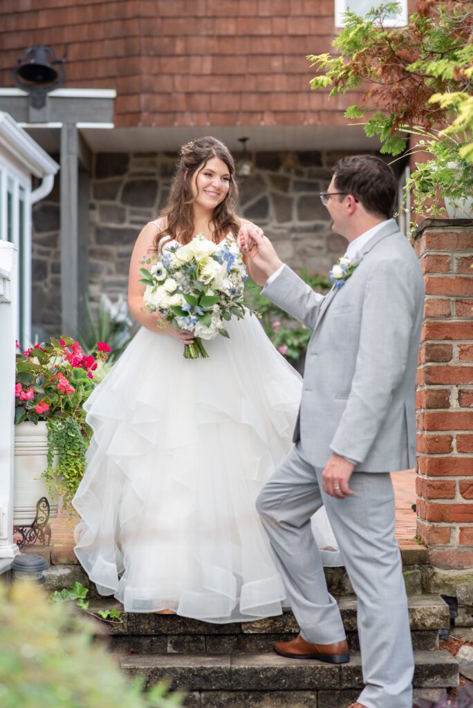 A bride in a white wedding gown holding a bouquet smiles at a groom in a gray suit as they stand on stone steps outside a building with flowers and greenery around them.