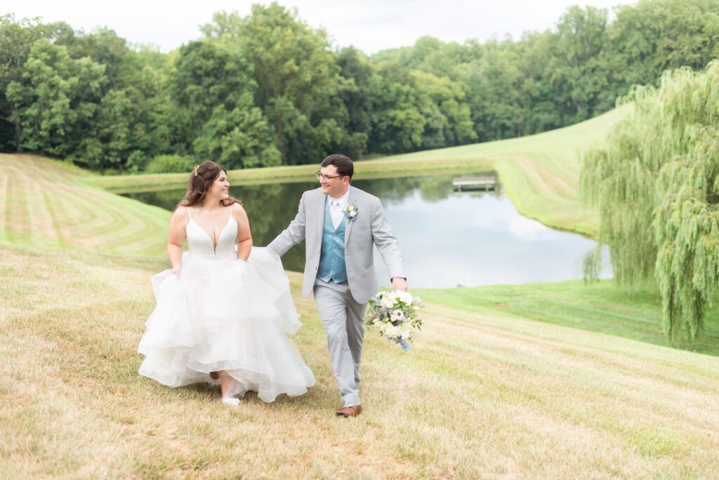 A bride in a white wedding dress and a groom in a gray suit and blue tie walk hand in hand across a grassy field with a pond and trees in the background on a cloudy day.