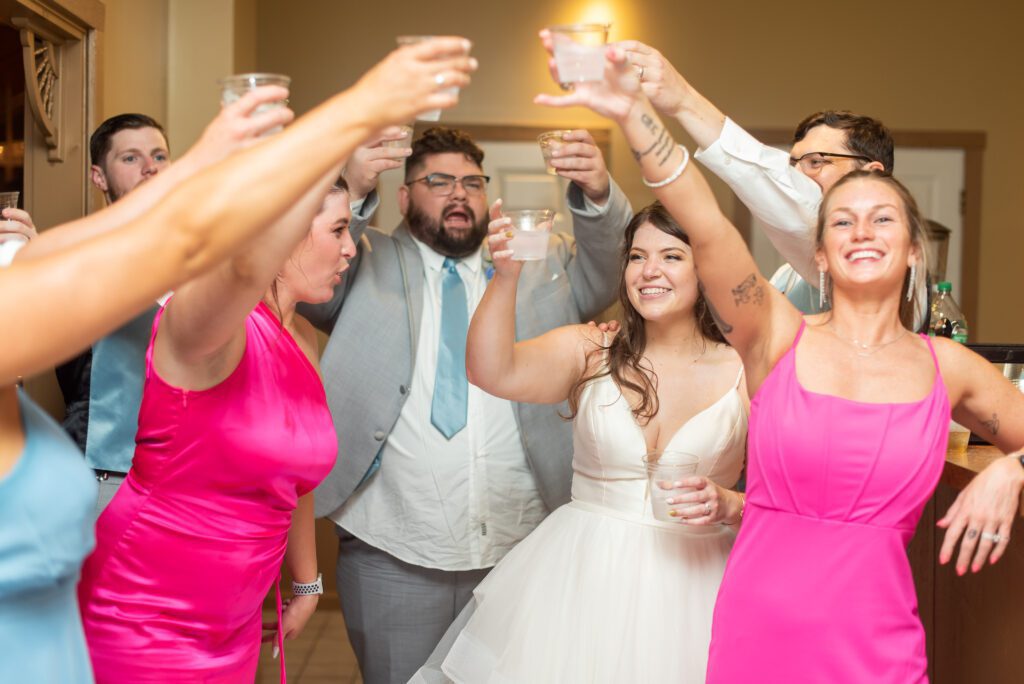 A group of smiling people raising their glasses for a toast at a celebration, with a bride in white at the center of the group.