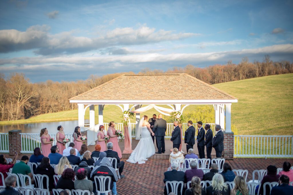 An outdoor wedding ceremony taking place under a white gazebo with a bride and groom at the altar, surrounded by groomsmen and bridesmaids, with seated guests facing them, all overlooking a scenic view of a pond and rolling hills under a partly cloudy sky.