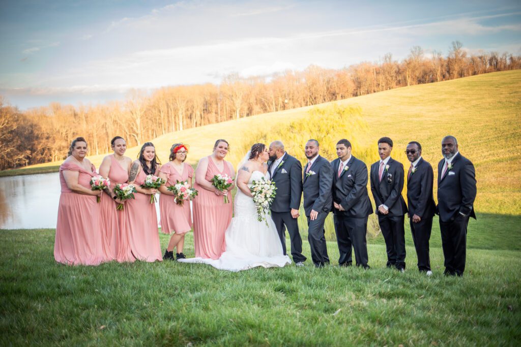 A wedding party posing outdoors with a bride and groom kissing, surrounded by bridesmaids in pink dresses and groomsmen in black suits, with a grassy field and a pond in the background under a clear sky.