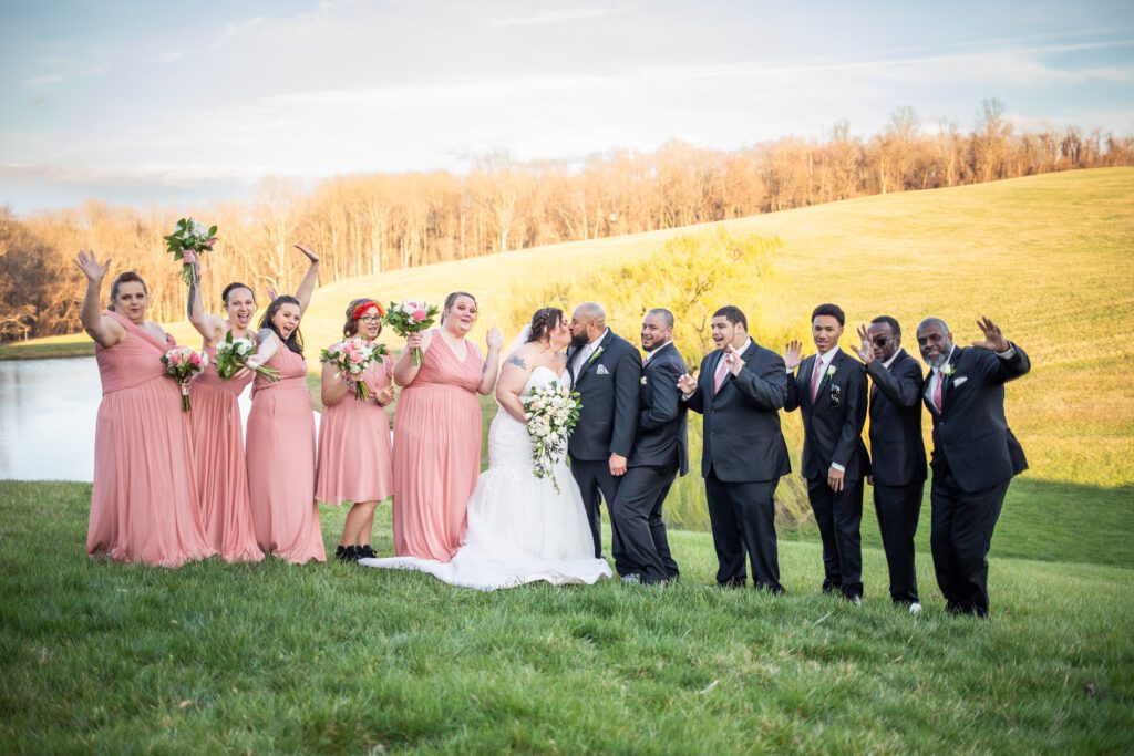 A wedding party posing for a photo outdoors with bridesmaids in pink dresses on the left, the bride and groom kissing in the center, and groomsmen in black suits on the right, all standing on a grassy hill with a lake and trees in the background.