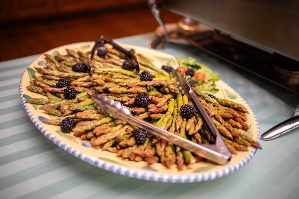 A large oval platter filled with roasted green asparagus spears topped with blackberries, flanked by tongs, on a striped tablecloth.