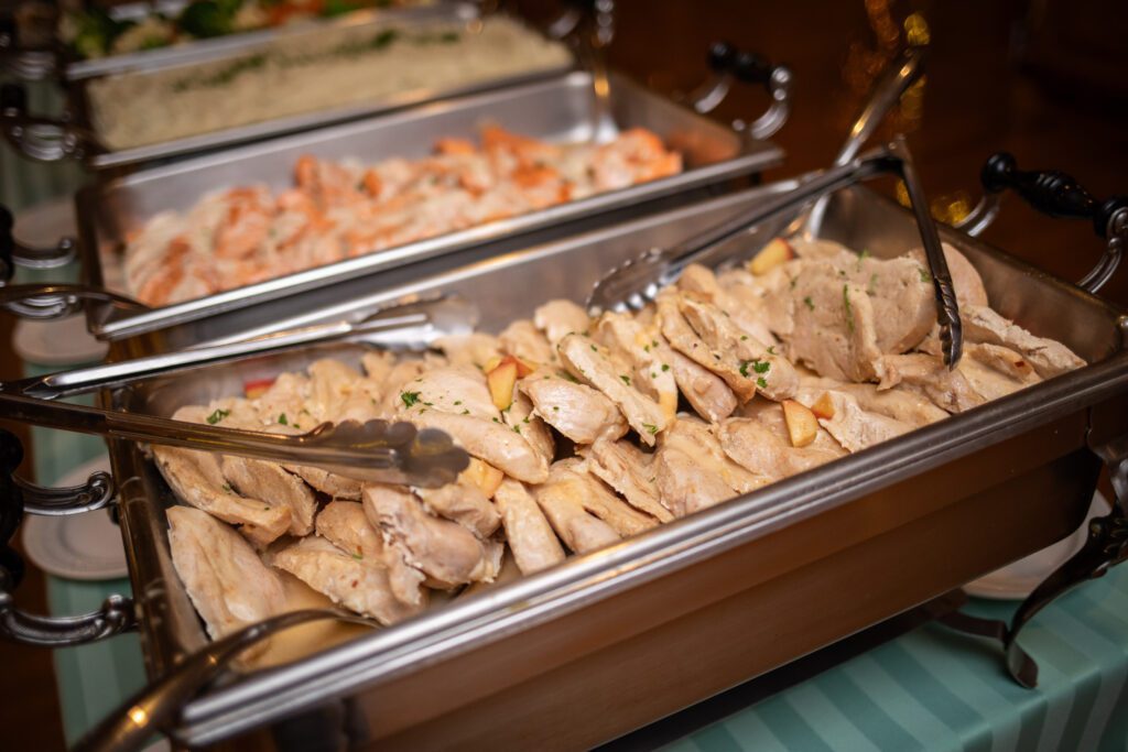 Buffet trays with gleaming serving utensils holding sliced chicken and shrimp dishes on a banquet table.