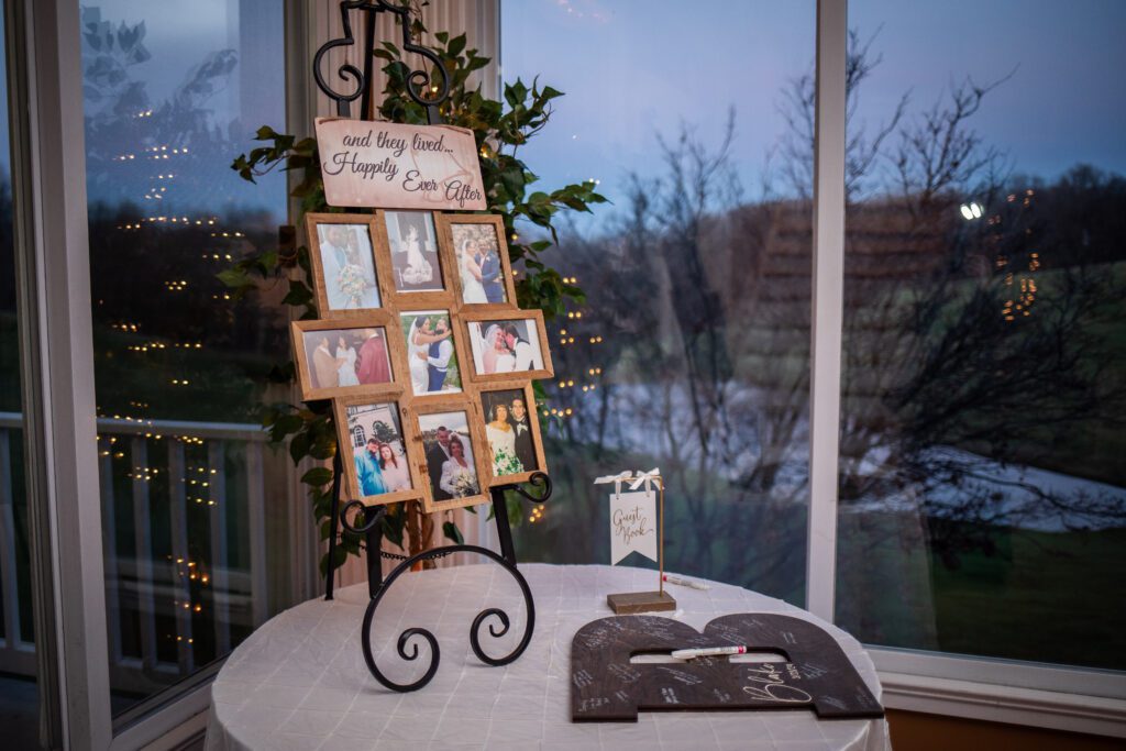 A decorated wedding photo display stand with the phrase "and they lived... Happily Ever After" amidst various framed couple photos, next to a guestbook table with a sign and markers, set against a window with evening outdoor lighting.