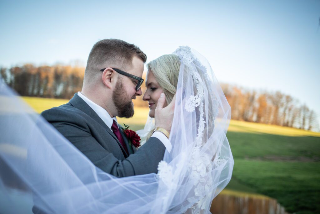 A bride and groom intimately facing each other with their foreheads touching, the bride's veil flowing in the foreground, with a backdrop of a field and trees under a clear sky.