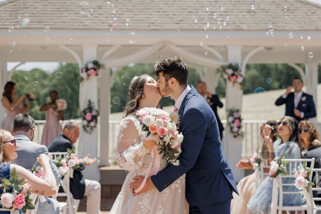 A newlywed couple shares a kiss at an outdoor wedding ceremony, standing under a gazebo adorned with floral decorations. The bride holds a bouquet of pink and white flowers. Guests, seated and standing around, applaud as bubbles float through the air. | Morningside Inn