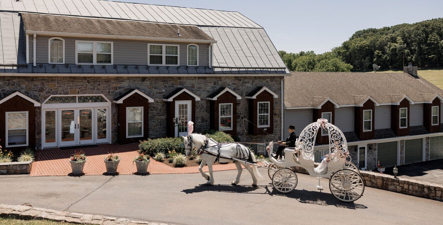 A white horse-drawn carriage carries a couple in wedding attire in front of a large stone and wood building with dormer windows, surrounded by potted plants and greenery on a clear day. | Morningside Inn