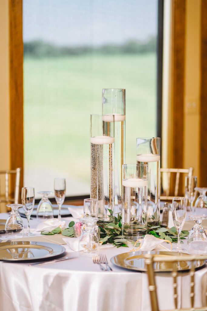 An elegantly set round dinner table adorned with a white tablecloth. The centerpiece features three tall glass vases with floating candles and greenery. Each place setting includes a plate, silverware, crystal glasses, and folded napkins, with a window view in the background. | Morningside Inn