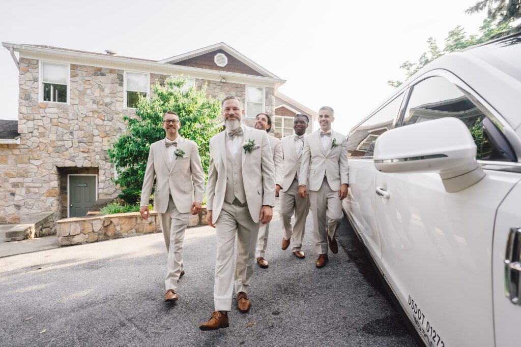 Five men wearing light-colored suits and brown shoes walk in a relaxed manner beside a white vehicle. They are in front of a stone house with greenery in the background, suggesting a wedding or formal event. | Morningside Inn