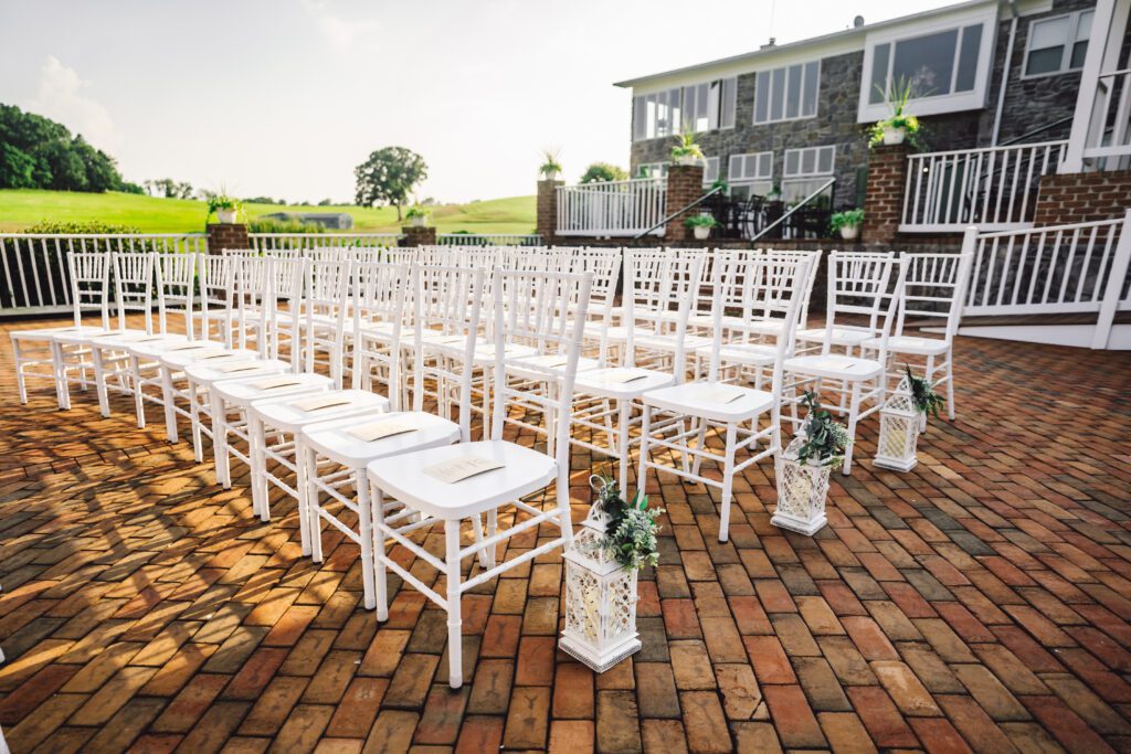 A brick-paved outdoor area is set up for a wedding with rows of white wooden chairs facing forward. Decorative lanterns with greenery hang from some chairs, with a large building and greenery visible in the background. Sunlight brightens the scene. | Morningside Inn