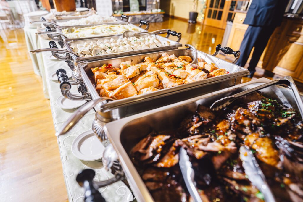 A buffet table filled with trays of various foods, including grilled meat, roasted vegetables, and pasta, with serving utensils. The setup is in an indoor setting with wooden floors and elegant decor, with a person partially visible on the right. | Morningside Inn