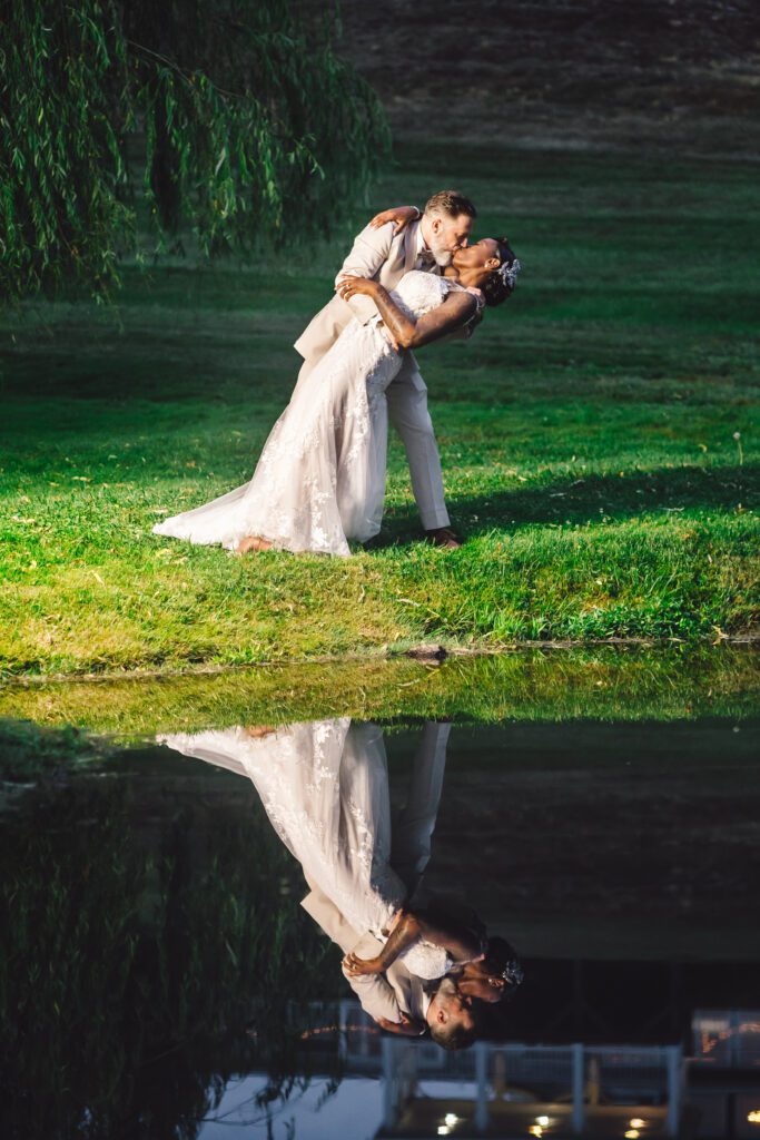 A bride and groom share a romantic kiss on a grassy area by a peaceful pond. The groom, dressed in a light suit, dips the bride, who is in a white lace dress. Their reflection is perfectly mirrored in the still water beneath them. | Morningside Inn