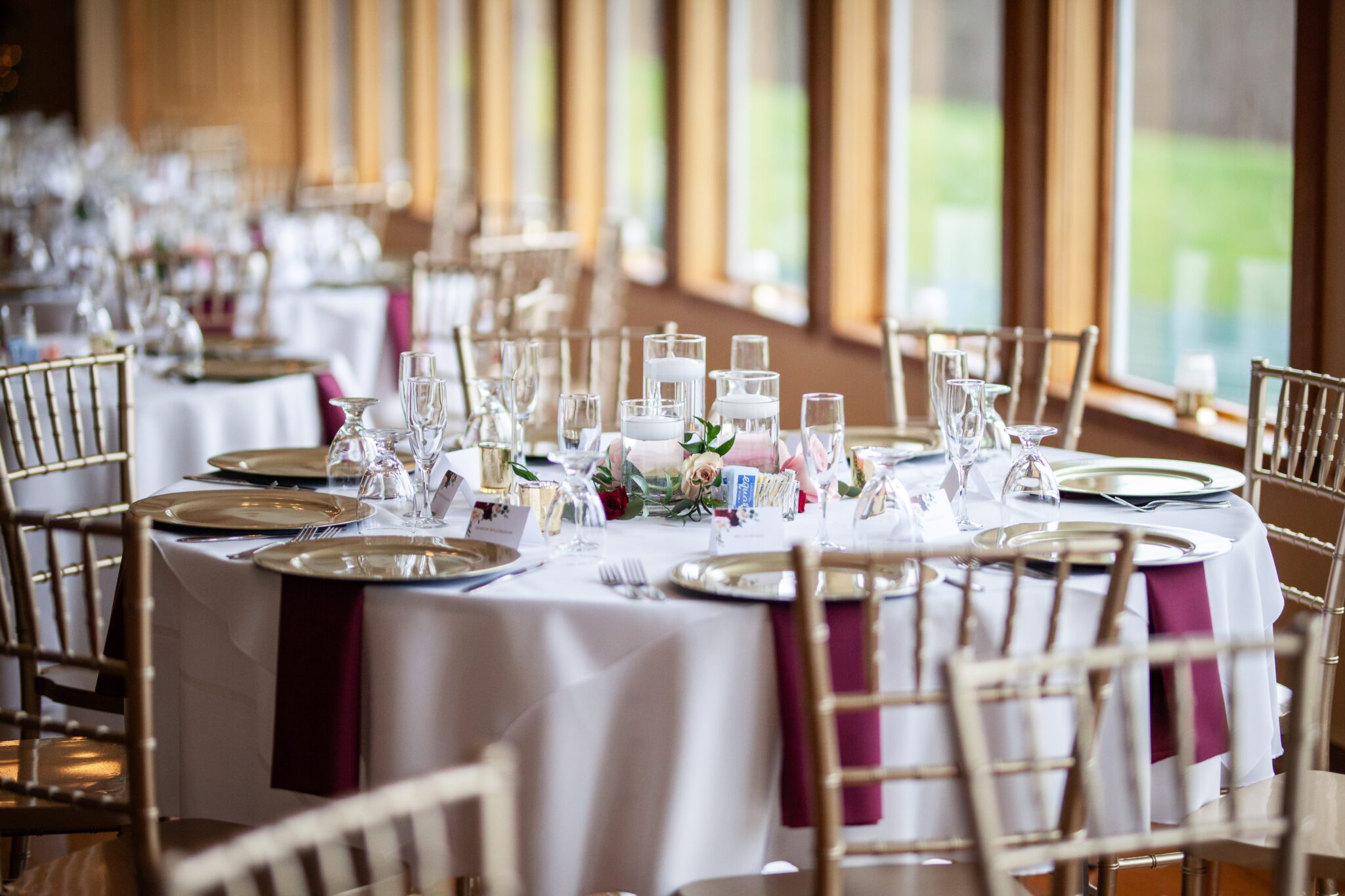 A beautifully set dining table with white tablecloths and elegant glassware in a bright, sunlit room at the Morningside Inn Wedding Venue. Each table is adorned with floral centerpieces and surrounded by gold chiavari chairs. Purple accents are visible among the settings. Large windows line one wall. | Morningside Inn