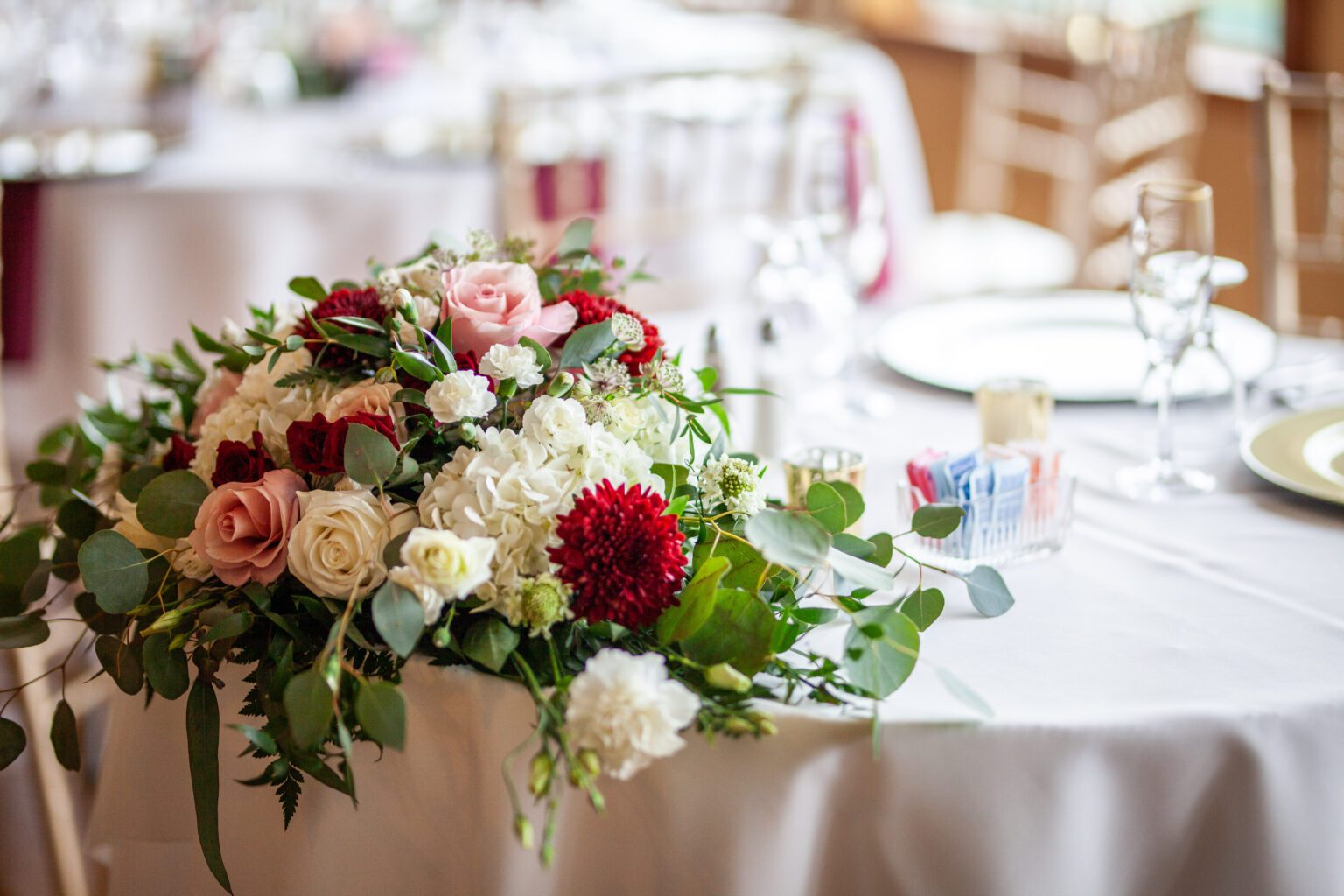 A beautifully decorated table at a wedding or formal event at the Morningside Inn. The table features an elegant floral arrangement with red and white carnations, pink and white roses, and lush greenery. In the background, other set tables and blurred chairs add to the charm captured in these wedding venue photos. | Morningside Inn