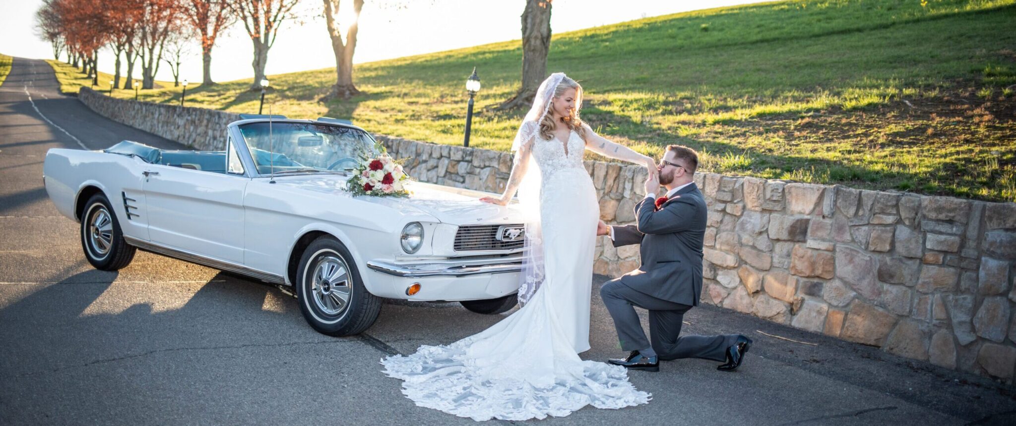 A bride in a white wedding gown and veil stands next to a white convertible with a bouquet on the hood. A groom in a grey suit kneels beside her, holding her hand. They are on a paved road with a stone wall and trees in the background. | Morningside Inn