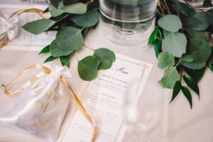 A wedding table setup at the venue features a menu card with elegant text, a small white gift bag tied with a gold ribbon labeled "thank you," and a glass vase with lush green eucalyptus leaves. The table is set with a white tablecloth, creating a sophisticated and natural ambiance for the ceremony. | Morningside Inn