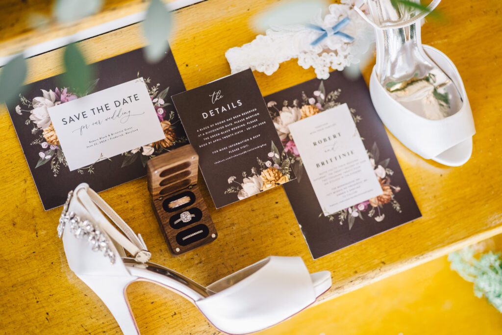 A flat lay of wedding items for an Elegant June Wedding at Morningside Inn, including white high-heeled shoes with a jeweled buckle, floral-themed 'Save the Date' and wedding detail cards, black and silver wedding rings, and a lace garter placed on a wooden surface. | Morningside Inn