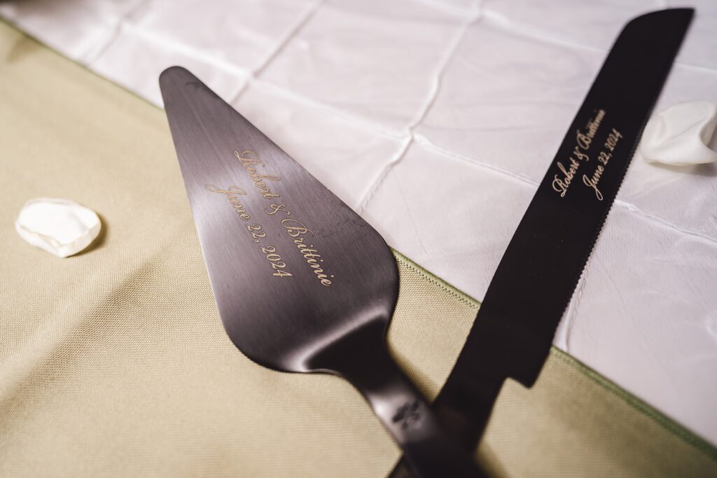 Close-up of a cake server and knife set engraved with the text "Brittinie & Robert, June 22, 2024." The tools rest on a soft green and white tablecloth with rose petals scattered around them, indicating an elegant June wedding at the Morningside Inn. | Morningside Inn