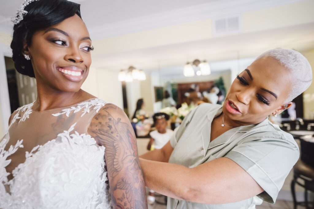 A bride with a tattoo on her arm smiles as she gets ready for her elegant June wedding at Morningside Inn. A woman in a green outfit adjusts the bride's dress, while reflections in the background show more people helping with preparations in a room filled with natural light. | Morningside Inn