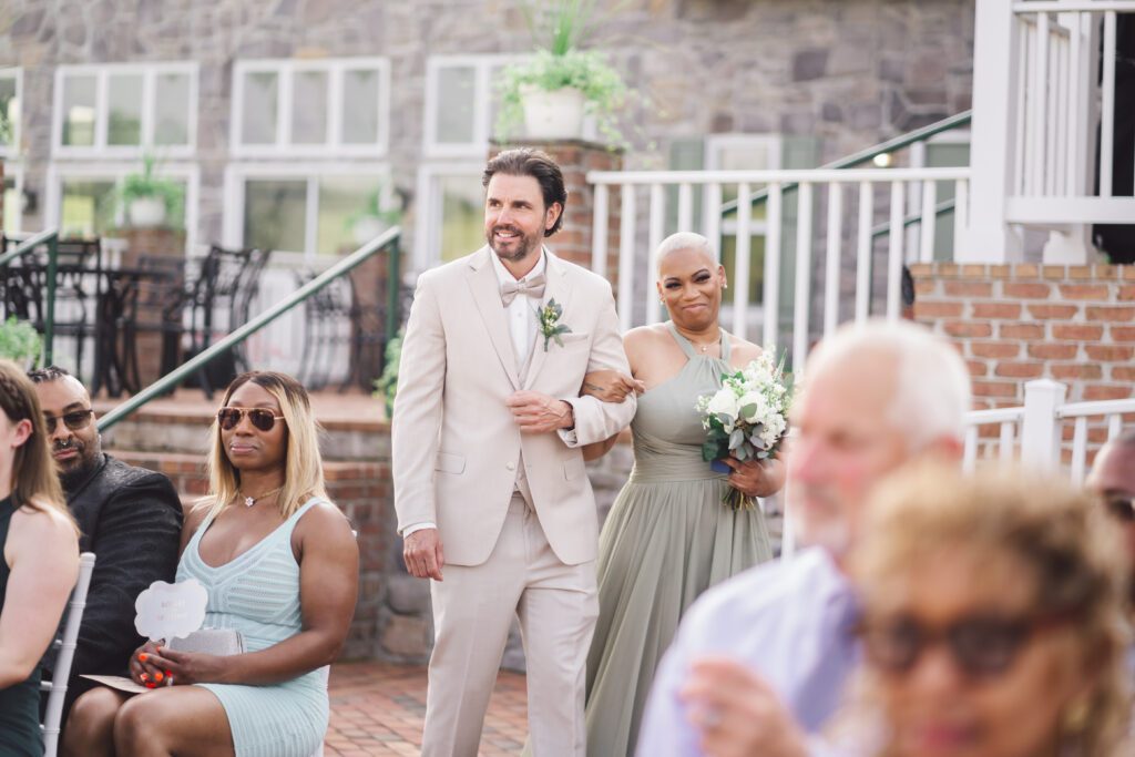 A man in a beige suit walks arm-in-arm with a woman in a light green dress, holding a bouquet of white flowers. They are outdoors at the elegant June wedding of Brittinie and Robert, with guests seated and watching them. The backdrop features the charming stone building and brick steps of Morningside Inn. | Morningside Inn