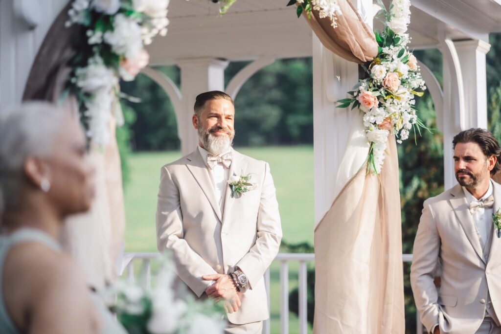 A groom stands under a decorated white arbor at Morningside Inn, smiling with his hands clasped in front of him. He is wearing a light suit with a bow tie and boutonniere. Another man in a similar outfit stands beside him. Floral arrangements and draped fabric adorn the space, perfect for an elegant June wedding. | Morningside Inn