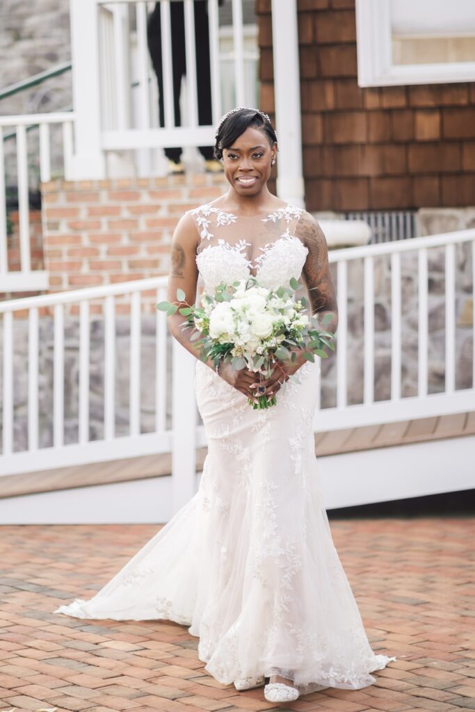 A bride is standing outdoors on a brick pathway at Morningside Inn, wearing a sleeveless, lace-embellished white wedding dress while holding a bouquet of white flowers with greenery. The background features a white rail and a building with brown shingles and brick detailing—an elegant June wedding setting. | Morningside Inn