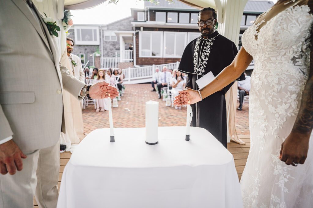 Brittinie and Robert, dressed in wedding attire, light a unity candle together during their elegant June wedding at Morningside Inn. The officiant stands behind the table with the candle as guests seated in the background watch the beautiful moment unfold. | Morningside Inn