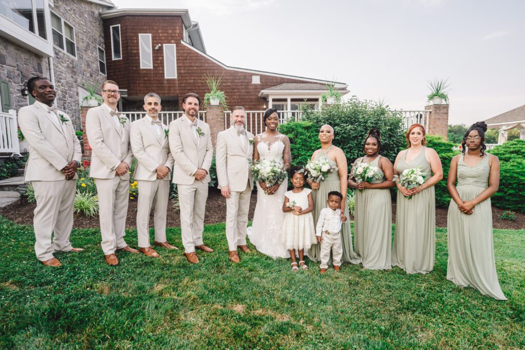 A wedding party stands outside with a house and lush greenery in the background. At the elegant June wedding at Morningside Inn, the bride and groom are centered, surrounded by bridesmaids in sage green dresses and groomsmen in beige suits. Two children, a boy and a girl, are also part of the group. | Morningside Inn