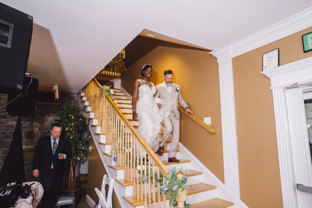 At Brittinnie and Robert's elegant June wedding at Morningside Inn, the bride in a white dress and the groom in a beige suit hold hands as they walk down a staircase adorned with greenery and flowers. The bride smiles at the guests while a man in a suit stands at the bottom of the stairs, seemingly preparing something. | Morningside Inn