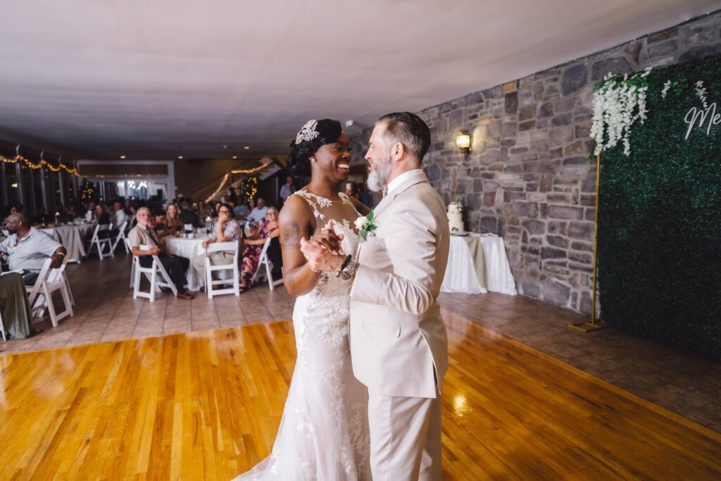 A bride and groom share their first dance at their elegant June wedding reception at Morningside Inn. The bride is wearing a white gown with intricate details, and the groom is in a light-colored suit. Guests are seated at round tables adorned with white linens, while string lights twinkle in the background. | Morningside Inn