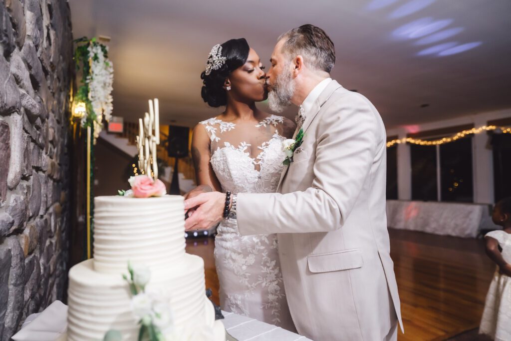 Brittinie and Robert share a kiss at their Elegant June Wedding next to a three-tiered white cake adorned with flowers. The bride wears a white lace gown, and the groom is in a light-colored suit. The backdrop features a stone wall and twinkling lights, reflecting the charm of Morningside Inn. | Morningside Inn