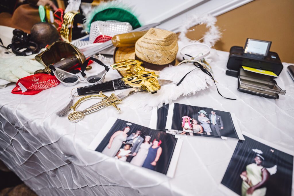 A cluttered table with various costume accessories including hats, oversized sunglasses, and feather boas sits ready for guests at an elegant June wedding at Morningside Inn. Several printed photos, a photo printer, and fun props are scattered across a white tablecloth, suggesting a festive photo booth setup. | Morningside Inn