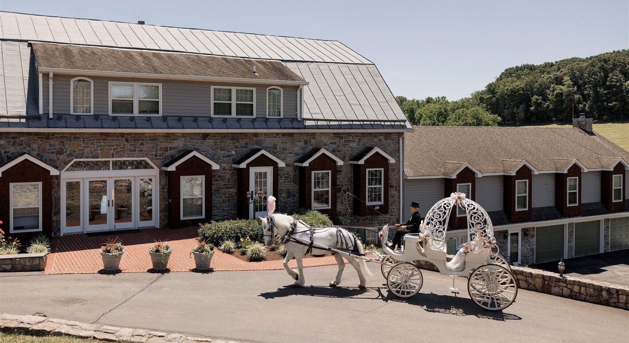 Wedding venue in Frederick Maryland with a white horse-drawn carriage with elaborate decorative designs is being pulled by a driver in formal attire in front of the top wedding venue in frederick maryland. Morningside Inn wedding venue features a stone facade and multiple peaked rooftop sections. The sky is clear and the surroundings are green. | Morningside Inn