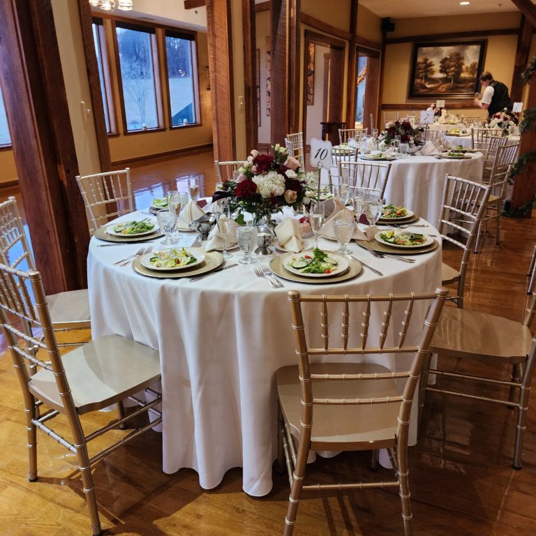 Elegant banquet hall set for a formal event with round tables dressed in white cloths, silver Chiavari chairs, and floral centerpieces. Each table is adorned with plated salads, rolled napkins, cutlery, glassware, and table number signs. Wood beams and a warm interior with paintings and windows add to the ambiance.