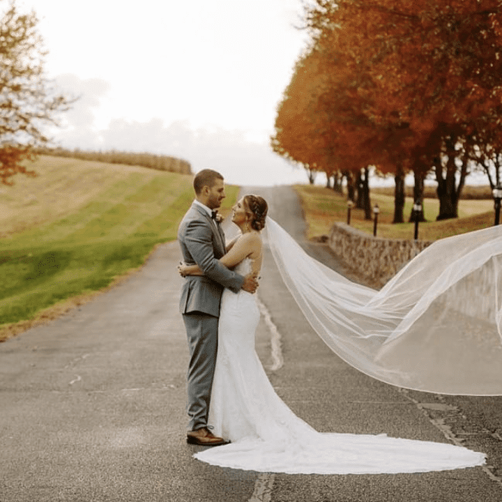 A bride and groom embracing each other on a pathway with the bride's veil flowing in the wind and autumn-colored trees in the background.